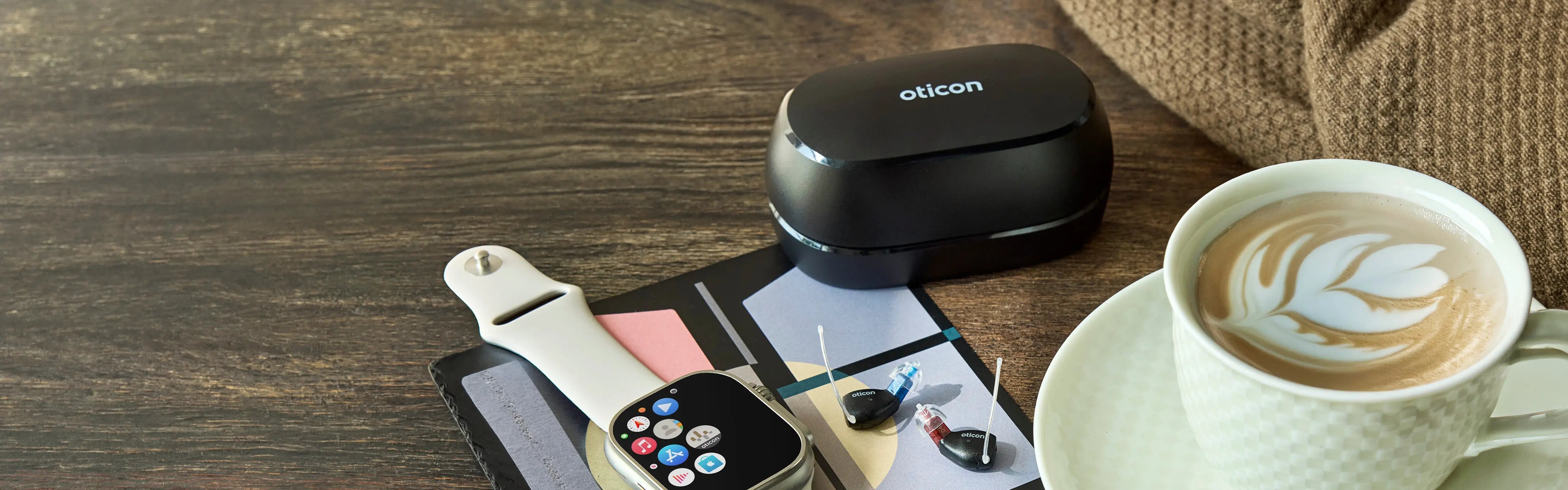 Oticon Zeal Hoergeraete praesentiert auf einem Holztisch mit einer Tasse Kaffee, einer Smartwatsch und Ladestation